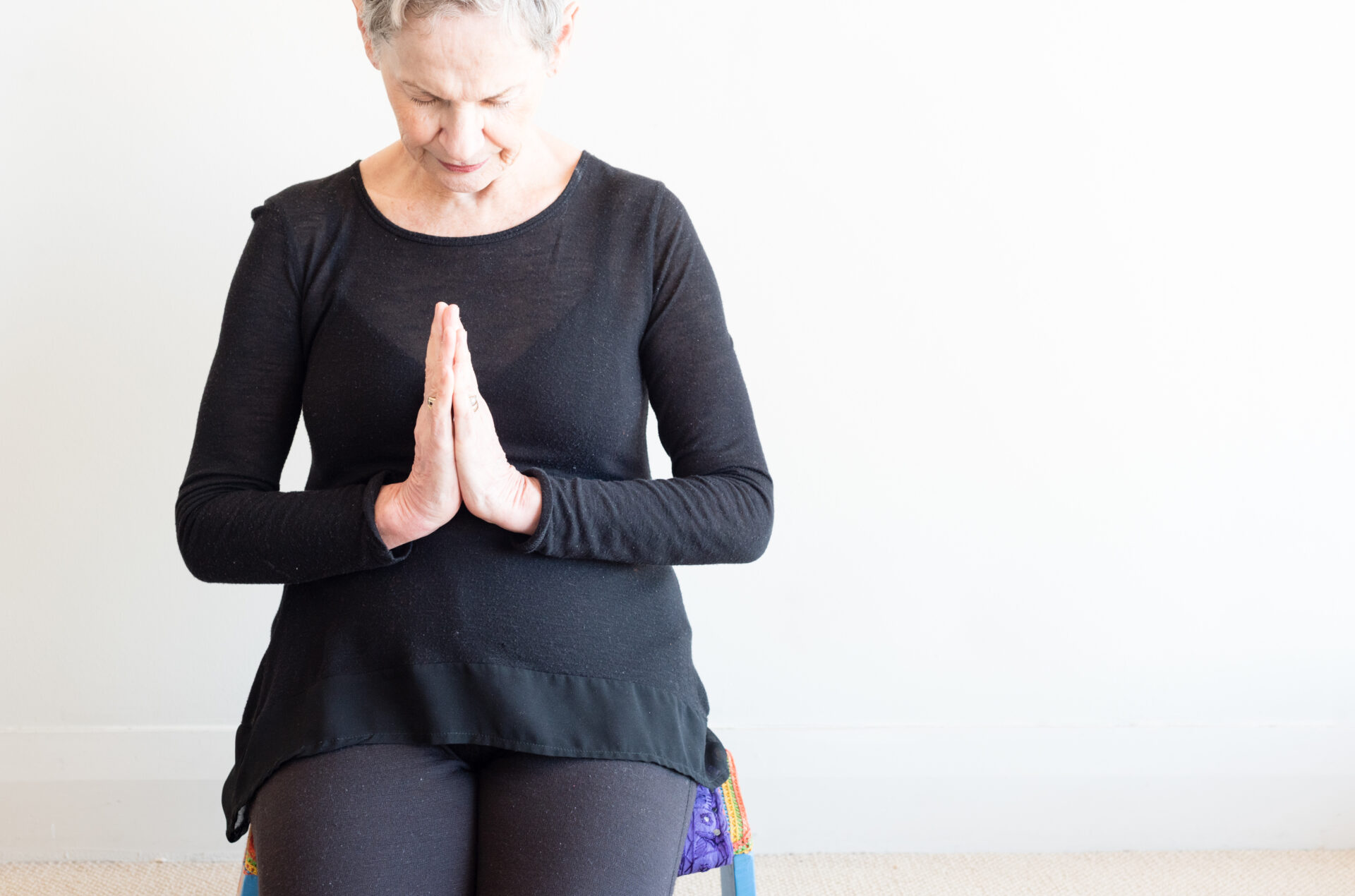 Older woman in prayer position Ausschnitt einer älteren Frau mit kurzen grauen Haaren und schwarzer Kleidung, die auf einem Yogastuhl sitzt und die Hände zum Gebet gefaltet hat.